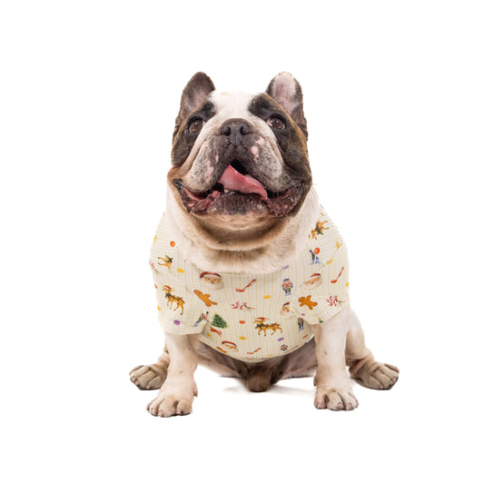 French Bulldog wearing a festive Christmas dog shirt with Santa, reindeer, nutcracker, candy cane, and holiday icons against a cream gingham background.