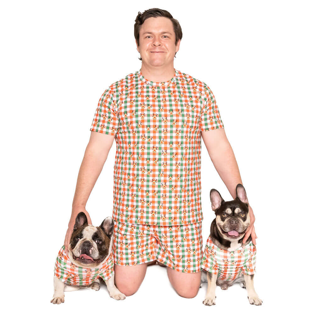 Two Ffrench Bulldogs wearing a Vibrant Hound Reindeer Christmas shirt for dogs. The shirt has green, red, and white cheekers with reindeer printed on it. A male is in the centre of the photo wearing a matching pyjama set with shorts and a T-Shirt.