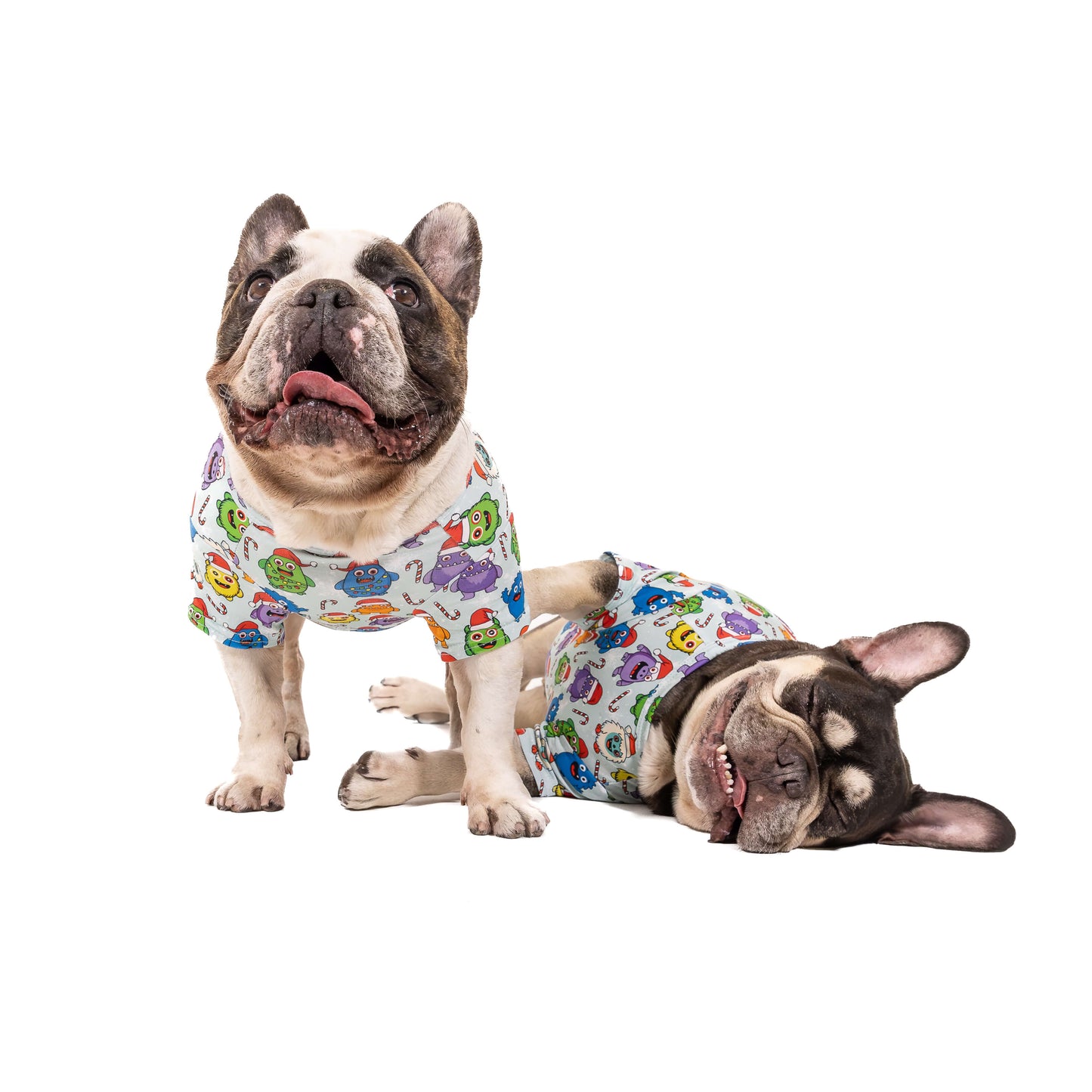 Two French Bulldogs wearing Festive Monsters Christmas dog shirts, one sitting and one lying down, both in colourful monster holiday print.
