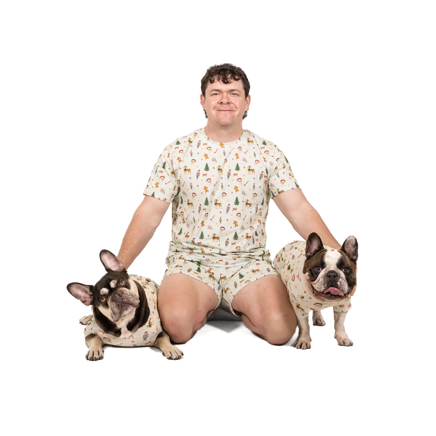 Man wearing Santa’s Workshop matching human Christmas pyjamas sitting with two French Bulldogs dressed in matching Christmas dog shirts.