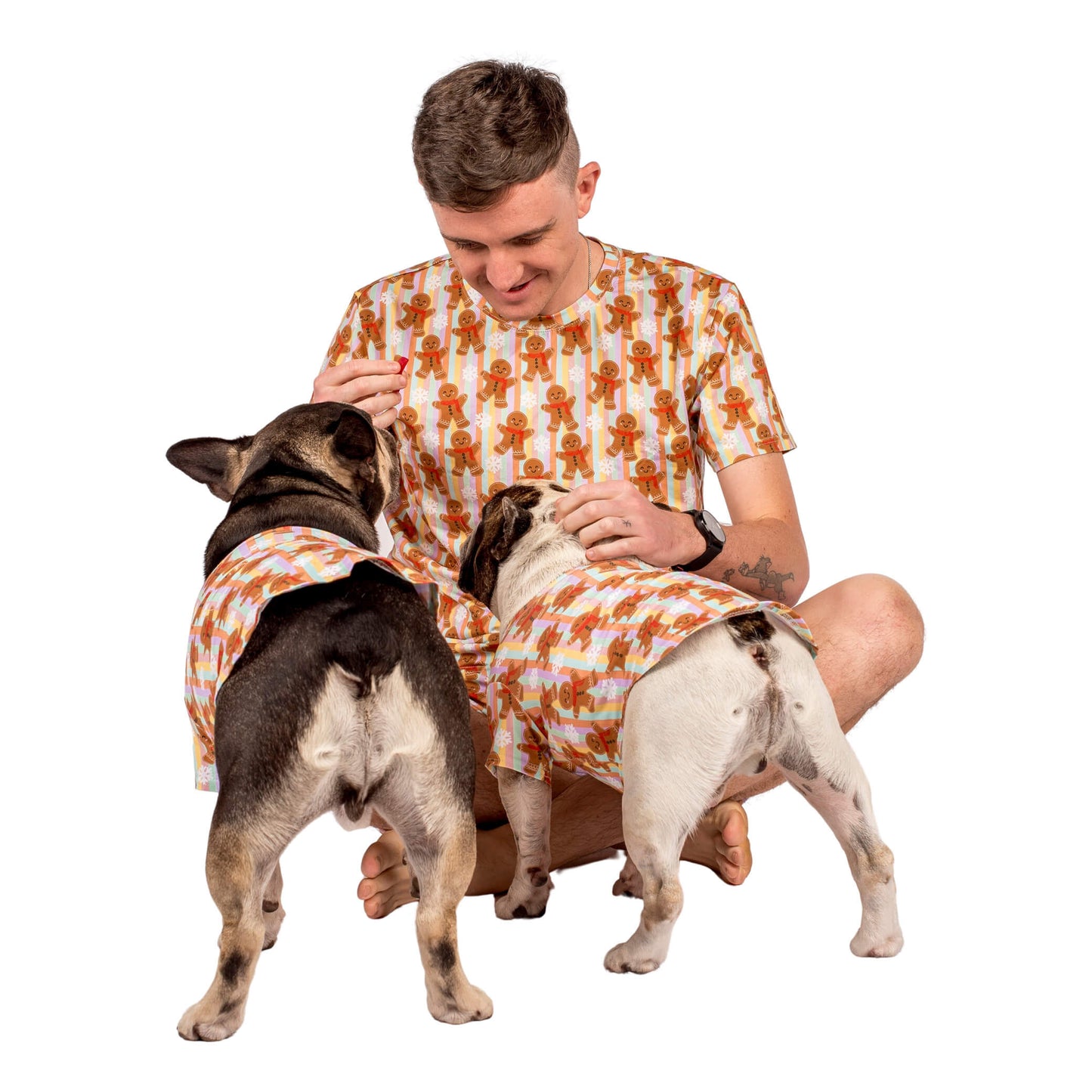 A dog dad sitting down cuddling two French Bulldogs. The human and dogs are wearing matching pyjamas. 